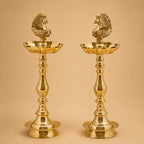 A pair of polished brass oil lamps with peacock figurines on top, set against a solid beige background.