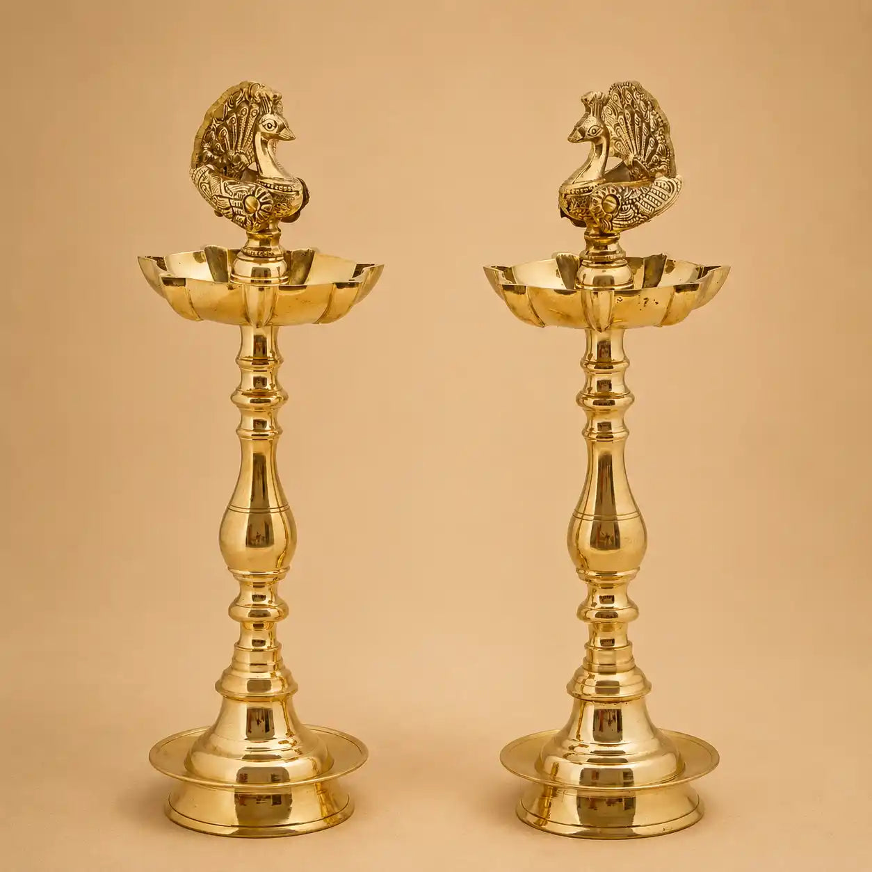 A pair of polished brass oil lamps with peacock figurines on top, set against a solid beige background.