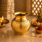 A polished brass pot with engraved floral patterns, placed on a marble surface with decorative props including a lit oil lamp, flowers, and a small bowl of red powder in a warm indoor setting.