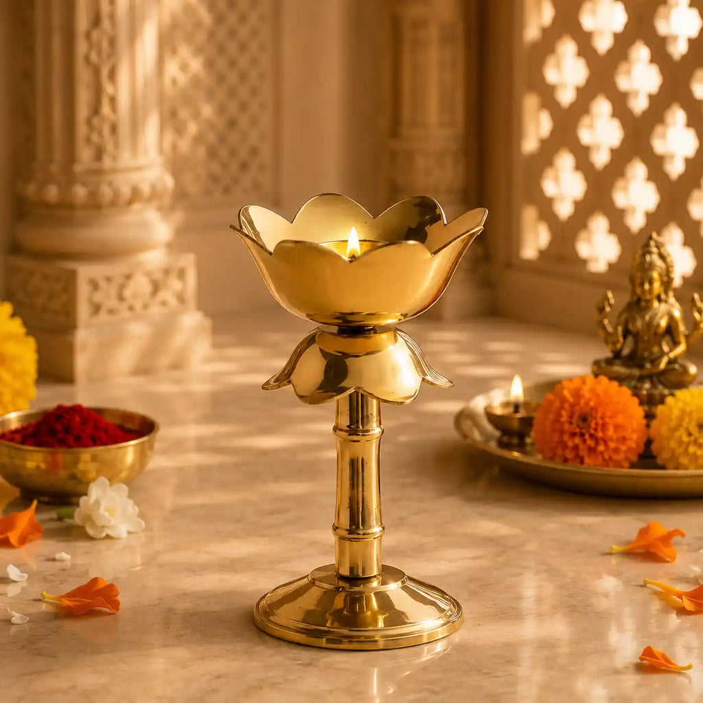 A polished brass lotus-shaped diya holder with a lit candle, set on a marble surface in a warm, decorated indoor setting with marigolds and a small brass statue.