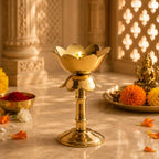 A polished brass lotus-shaped diya holder with a lit candle, set on a marble surface in a warm, decorated indoor setting with marigolds and a small brass statue.