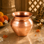 A polished copper pot (kumkum pot) with a flared rim, set on a stone surface surrounded by marigold flowers, a lit brass diya, and a brass Ganesha idol in a warm, traditional setting.