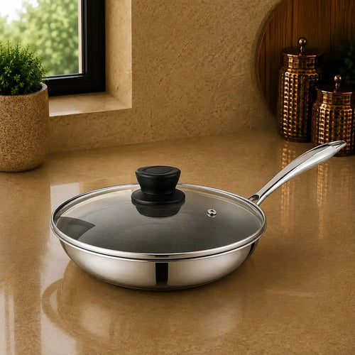 A stainless steel sauté pan with a clear glass lid and black knob, sitting on a beige countertop in a kitchen setting.