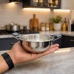 A hand holds a small stainless steel pot with black handles in a modern kitchen setting.