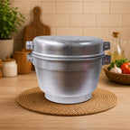 A large, round, double-walled stainless steel pot or steamer pot with side handles, sitting on a round woven jute placemat on a wooden countertop in a kitchen setting.