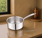 A stainless steel saucepan with a long handle sits on a polished countertop in a kitchen setting, with decorative copper canisters and a potted plant in the background.