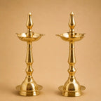 Two traditional brass oil lamps with ornate fluted stems and pointed finials, shown side by side against a warm beige background.