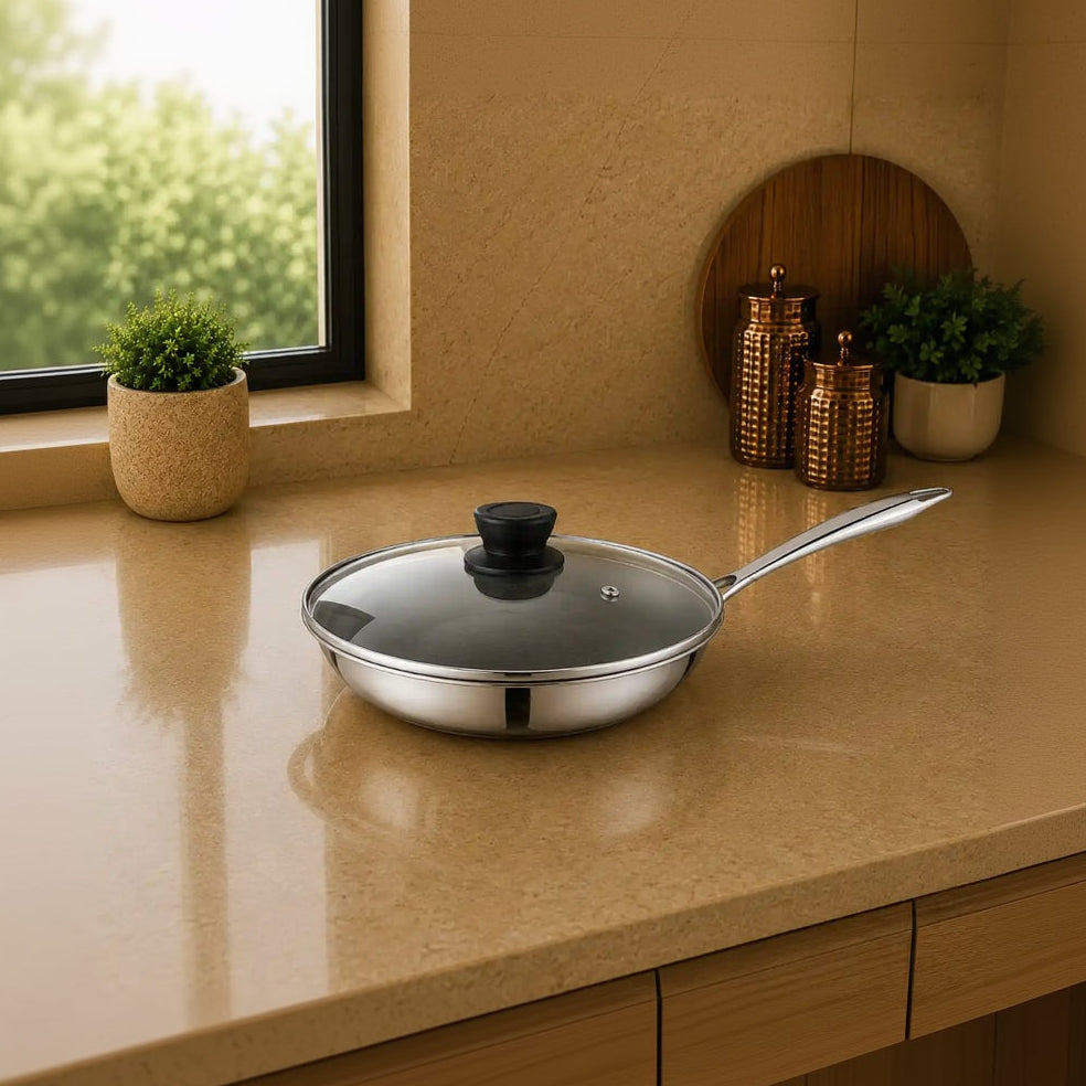 A stainless steel frying pan with a clear glass lid and black knob sits on a beige kitchen countertop near a window, with decorative plants and canisters in the background.