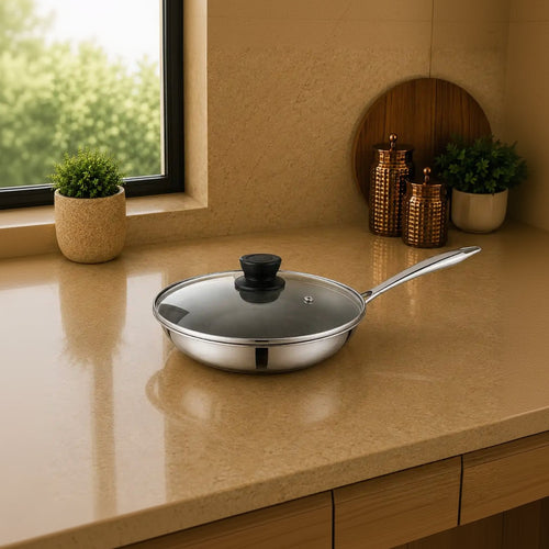 A stainless steel frying pan with a clear glass lid and black knob sits on a beige kitchen countertop near a window, with decorative plants and canisters in the background.